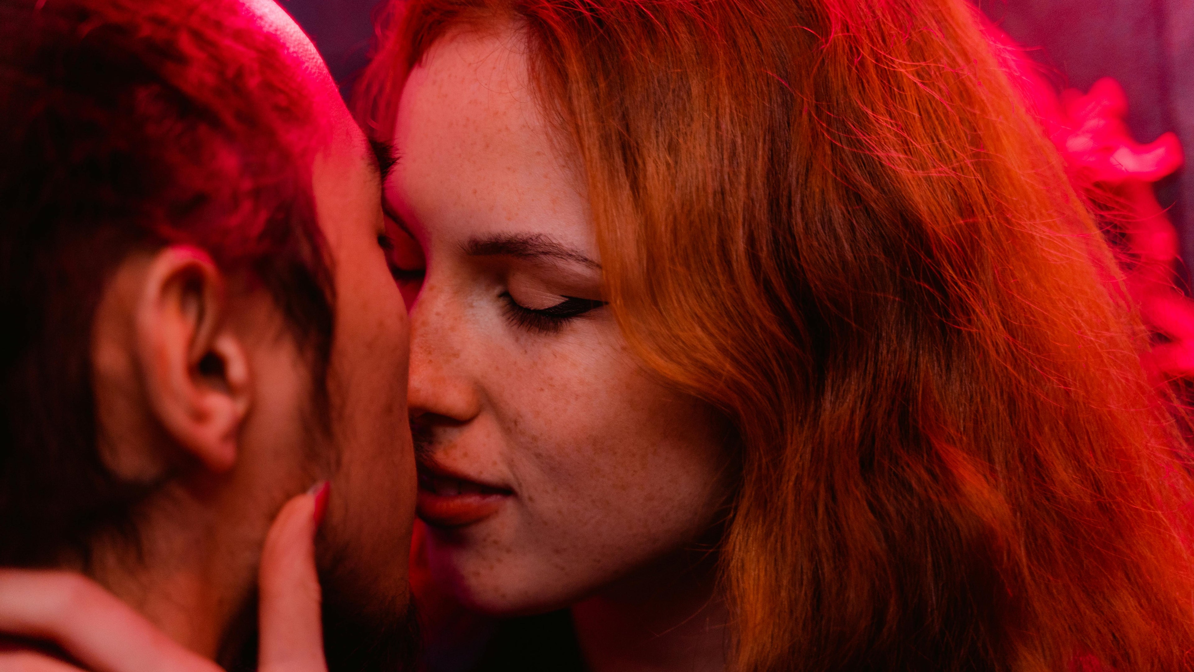 Two people with red hair in a close, intimate pose under red lighting.
